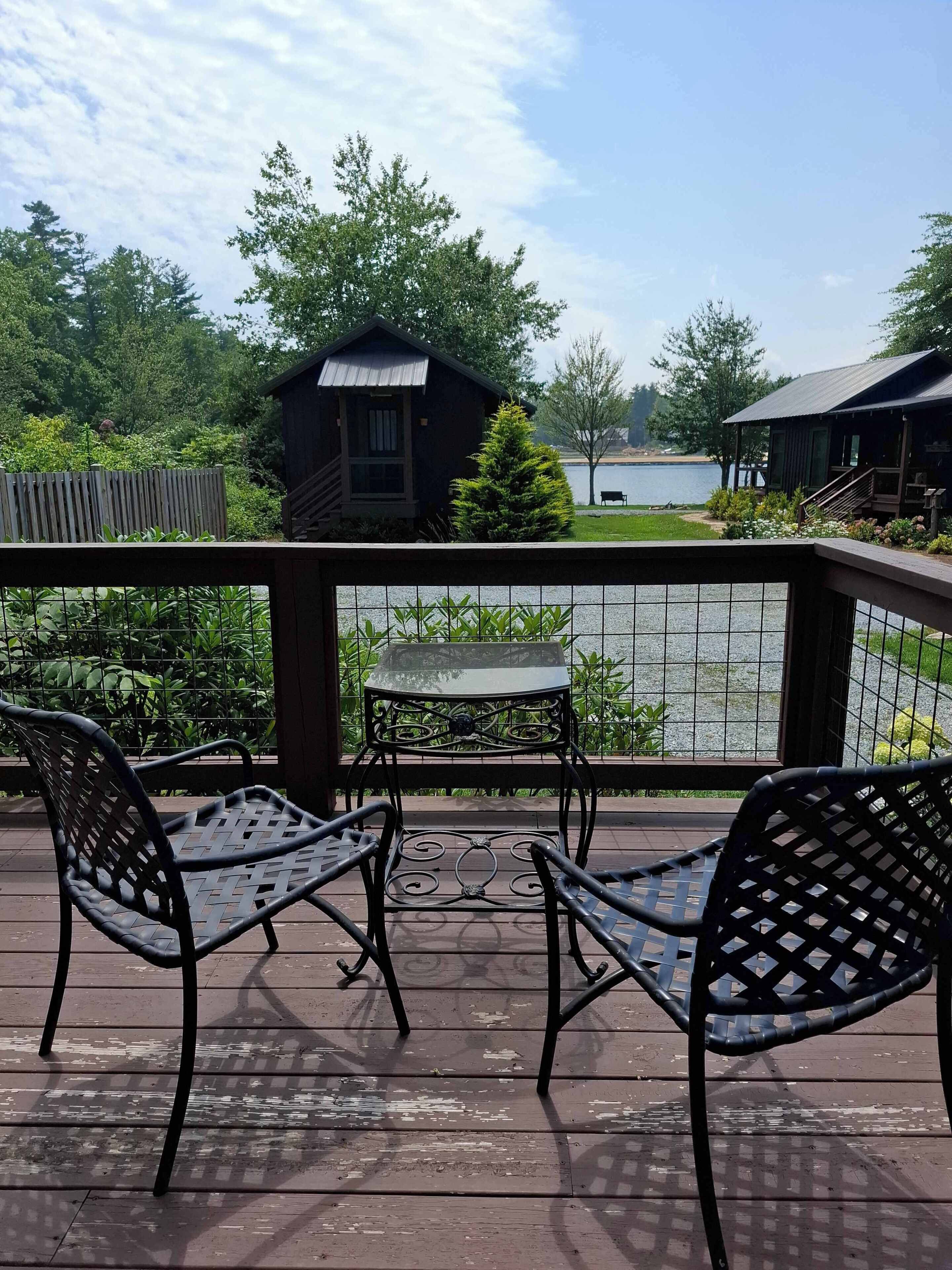 A wooden deck with two metal chairs and a small table, overlooking a lake with a wooden cabin and a dock in the background.