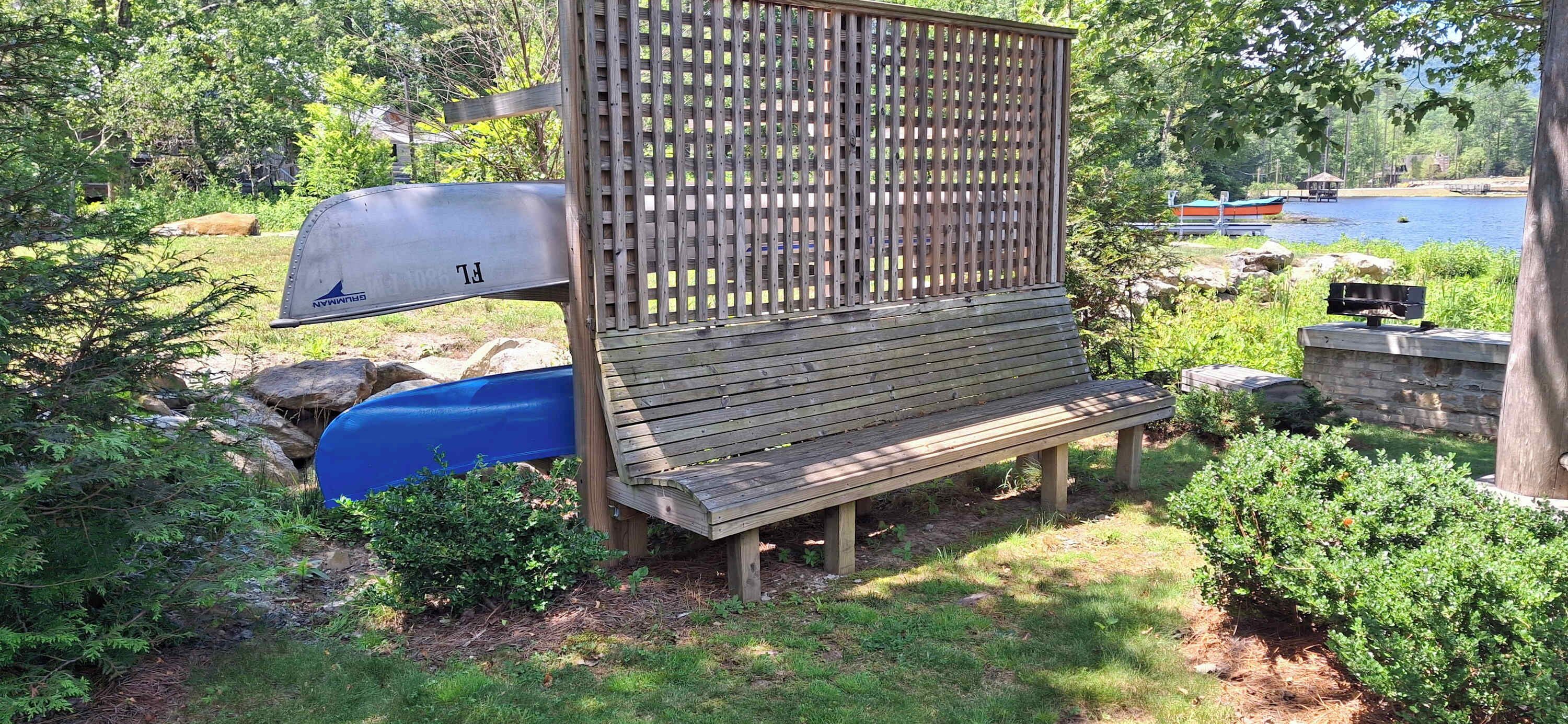 A wooden bench with a metal grill attached to a wooden structure, surrounded by greenery and a body of water in the background.