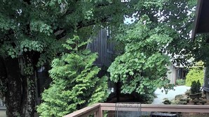 A wooden deck with a metal table and chairs, surrounded by lush greenery and a building in the background. - The Cedar Cabin | Lake Views, Walk to Cashiers (Cashiers)