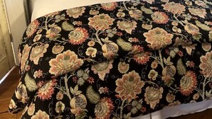 A bed with a floral-patterned bedspread, a white pillow, and a red accent pillow. There are two wall-mounted lamps with black shades and a rustic wooden headboard. - The Cedar Cabin | Lake Views, Walk to Cashiers (Cashiers)