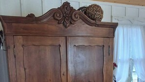 A wooden wardrobe with intricate carvings, a window with sheer curtains, and a patterned cushion on a wooden surface. - The Cedar Cabin | Lake Views, Walk to Cashiers (Cashiers)
