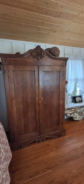 Un armario de madera con tallas intrincadas, una ventana con cortinas translúcidas y un cojín estampado sobre una superficie de madera.