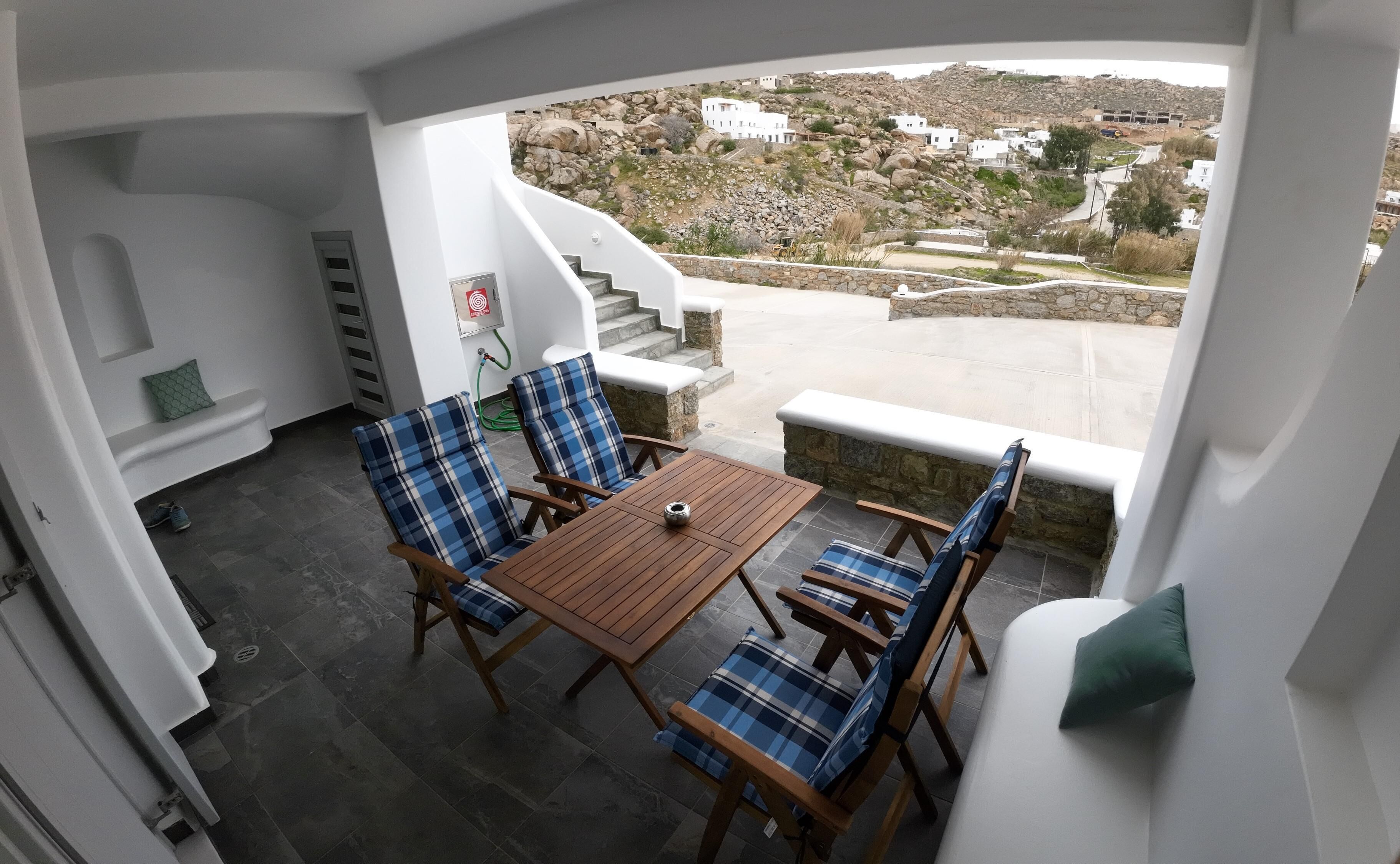 Ein Balkon mit einem Holztisch und blauen karierten Stühlen, mit Blick auf eine felsige Landschaft mit weißen Gebäuden.