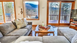 Un salón con un sofá gris, una mesa de centro con libros y un ventanal con vistas a la montaña.