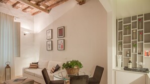 Een woonkamer met een glazen salontafel, een bank en een stoel. Er hangen fotolijstjes aan de muur en een plank met flessen en glazen.