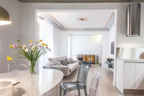 A modern living room with a marble dining table, clear chairs, a sofa, and a kitchen area with white cabinets and a stainless steel range hood.
