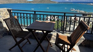 Ein Balkon mit zwei Holzstühlen und einem kleinen Tisch, mit Blick auf eine Küstenlandschaft mit Booten und Bergen im Hintergrund.