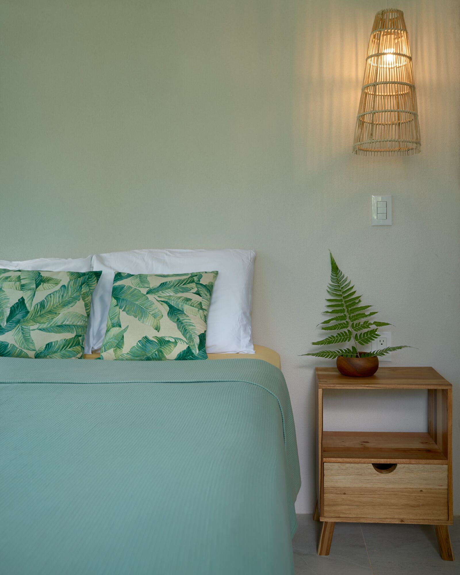 Uma cama com colcha verde e duas almofadas, uma mesinha de cabeceira com planta, e luminária fixada na parede.