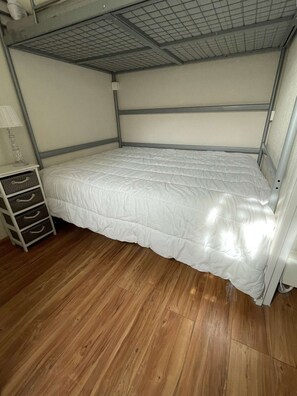 Un lit superposé avec un matelas blanc et une structure en métal, un plancher en bois et une petite table de chevet avec des tiroirs.