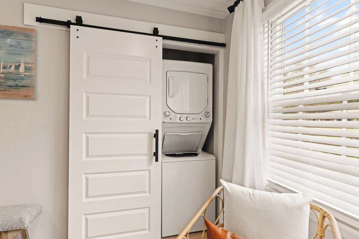 A room with a sliding door leading to a laundry area, a wicker chair with a cushion, and a window with blinds.