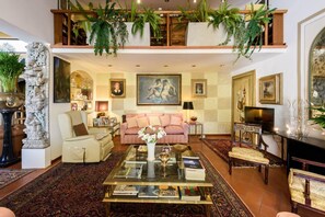 A spacious living room with a high ceiling, a sofa set, a coffee table with books, a fireplace, and a large painting.