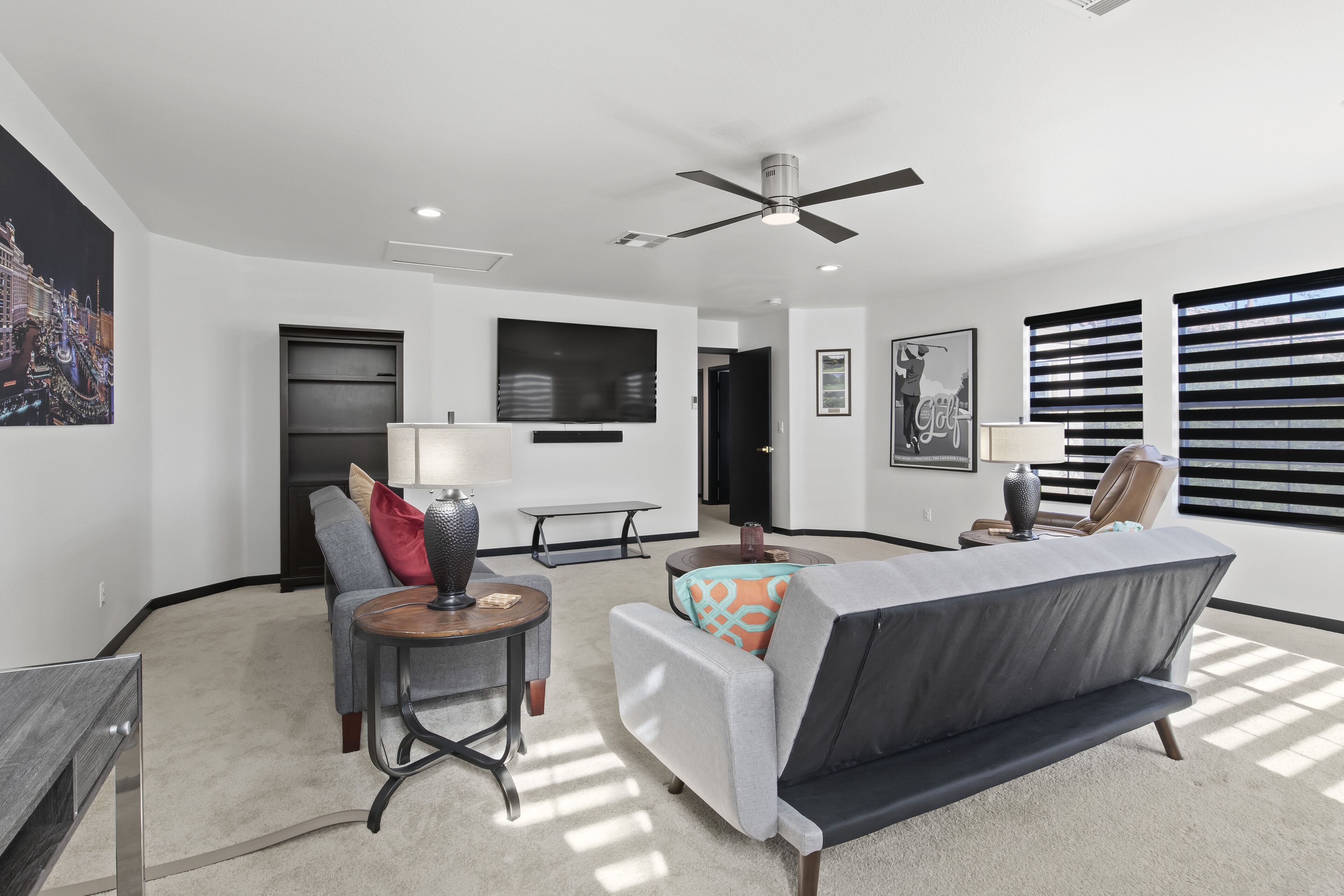 A modern living room with a grey sofa, a round coffee table, and a TV. There is a ceiling fan, a painting on the wall, and a window with blinds.