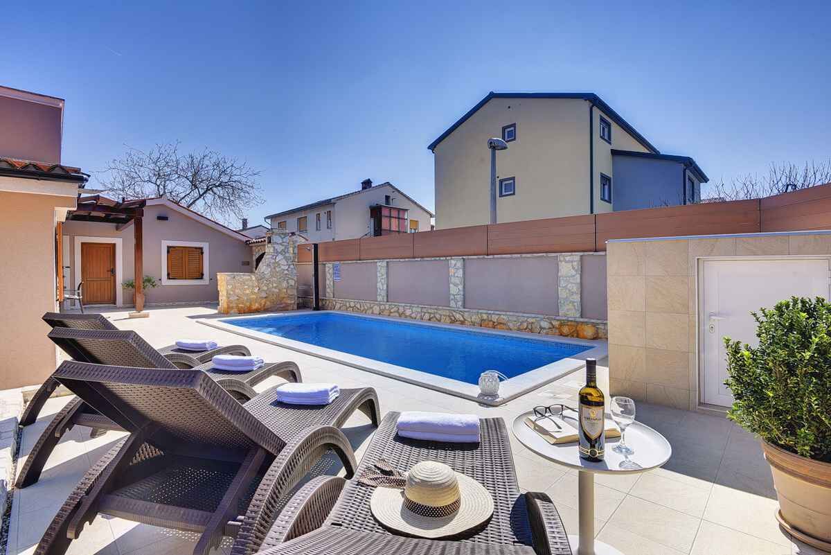 A swimming pool with lounge chairs and a table, surrounded by a low stone wall and a building.