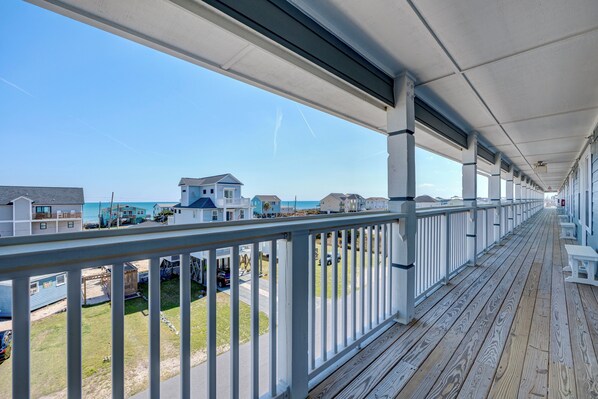 Une terrasse en bois couverte, dotée de rampes blanches, qui offre une vue sur une plage et des maisons.