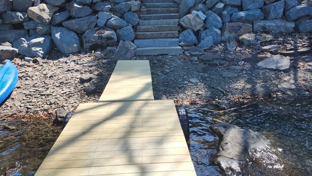 A wooden bridge over a rocky stream with a stone wall on one side and a house in the background.