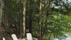 A lakeside campsite with Adirondack chairs, a fire pit, and a gravel path.
