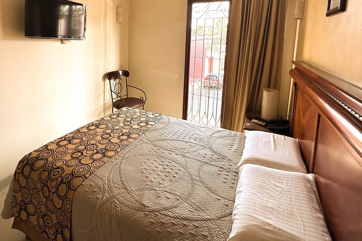 A hotel room with a bed, a chair, a television mounted on the wall, and a window with curtains.