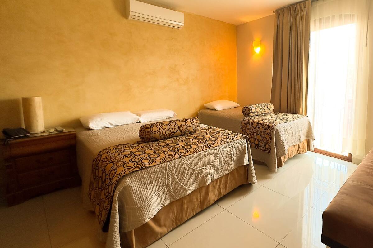 A hotel room with two beds, a bedside table with a lamp, a window with curtains, and a wall-mounted air conditioner.
