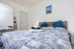 Een slaapkamer met een bed, kussens, een nachtkastje, een dressoir, een deur en een lijst met een afbeelding aan de muur.