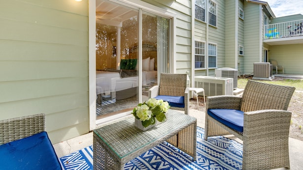 Un patio exterior con mobiliario de mimbre, una mesa con superficie de vidrio y un florero, y un área cubierta con vista a la casa.