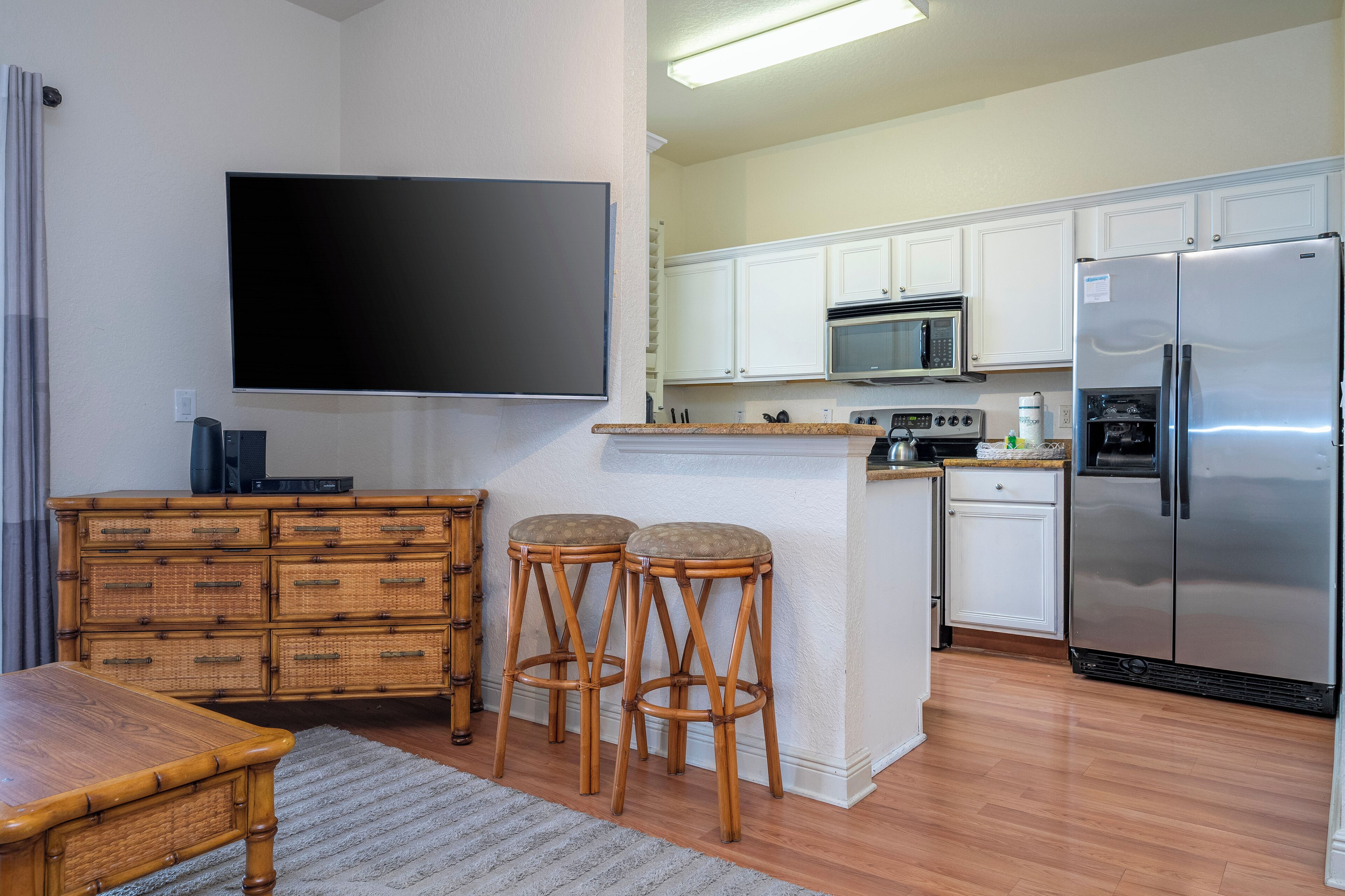 A modern kitchen with white cabinets, a stainless steel refrigerator, a microwave, and a flat-screen TV mounted on the wall. There are two wooden stools and a dining table with a light-colored rug.