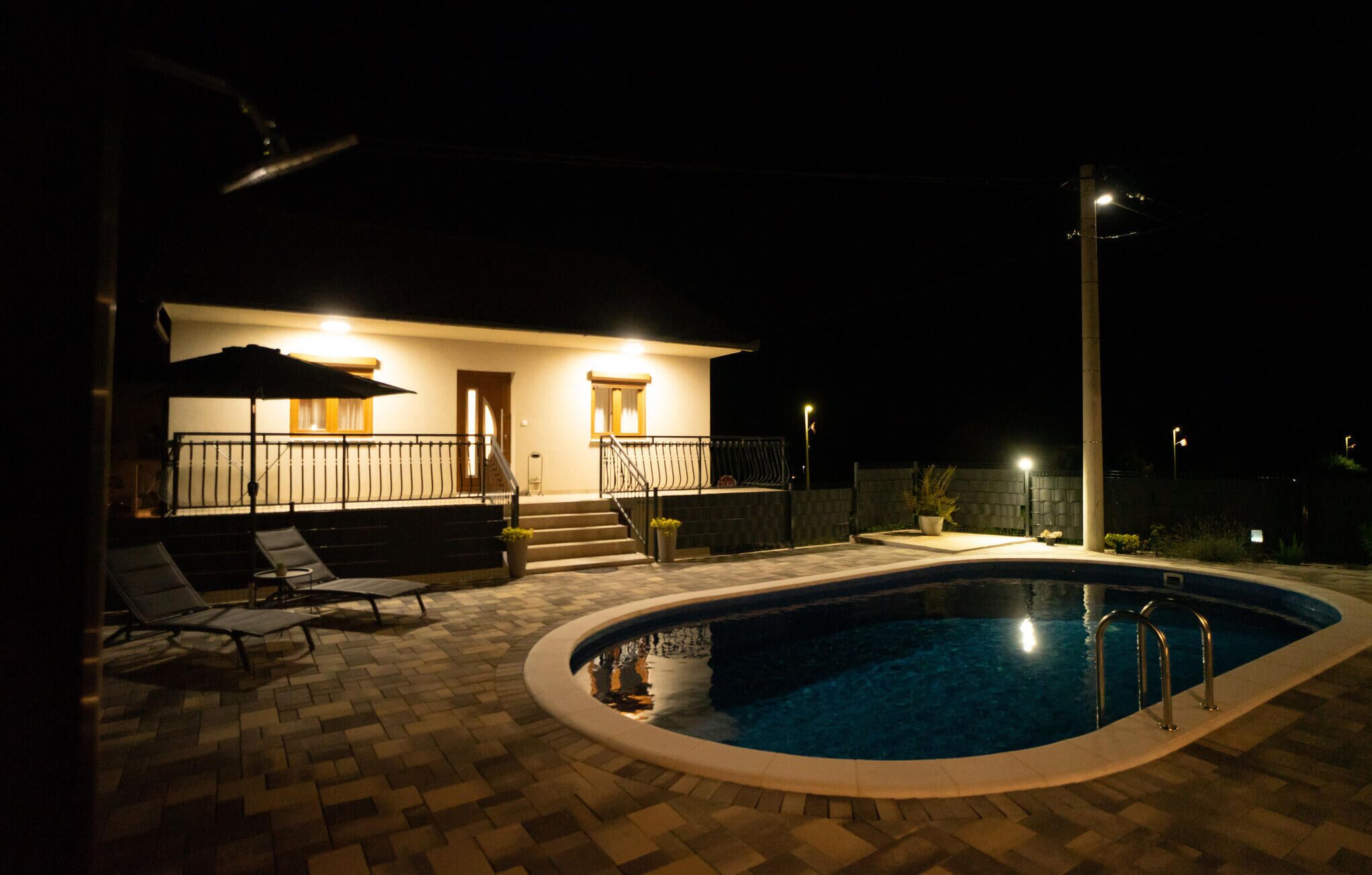 Ett poolområde som är vackert upplyst på kvällen, med ett hus, utomhusmöbler och en välbelyst swimmingpool.