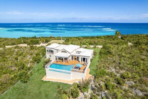 A large house with a swimming pool, surrounded by lush greenery and overlooking a clear blue ocean.