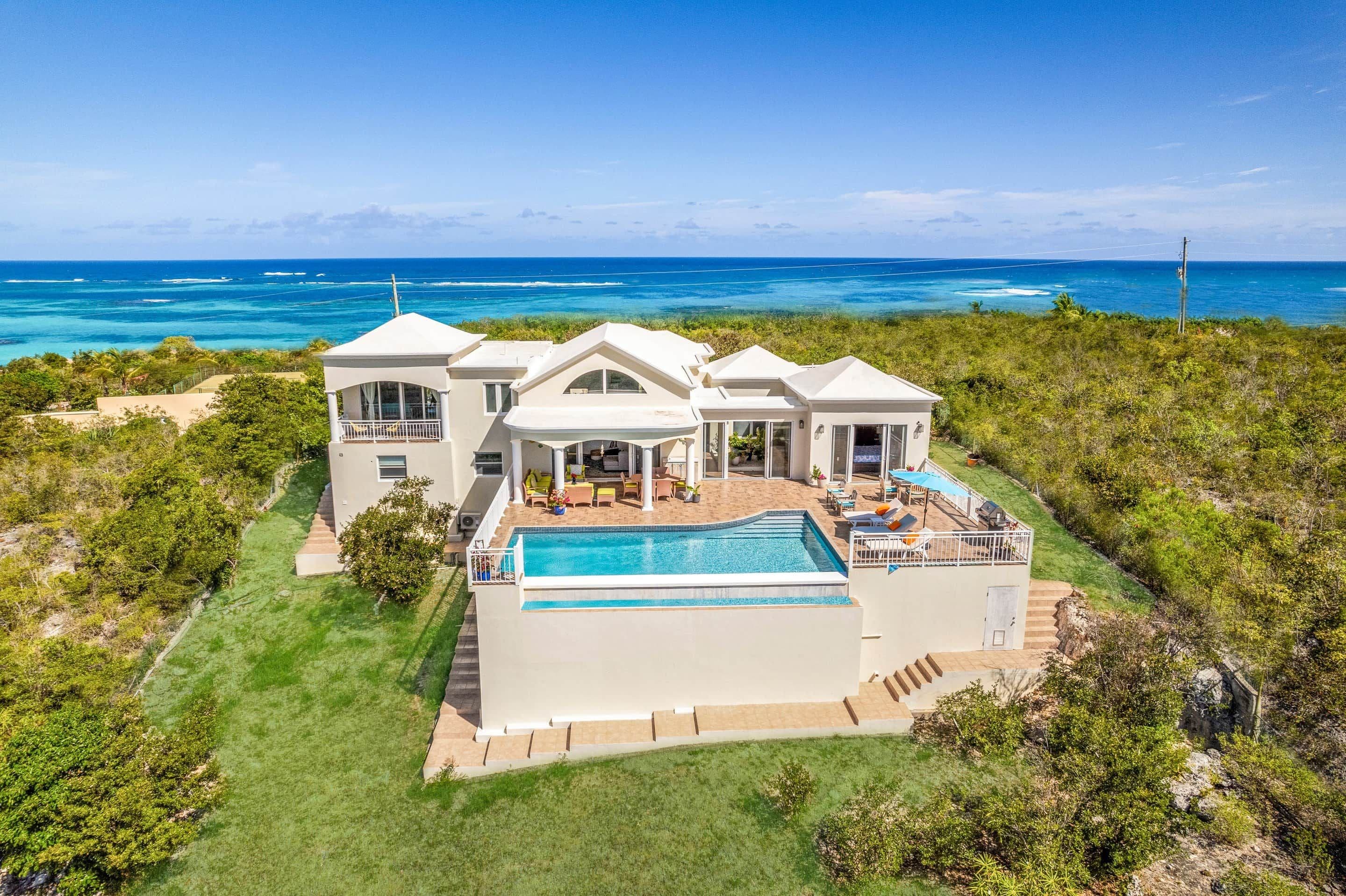 A large, modern house with a swimming pool, surrounded by lush greenery and overlooking a clear blue ocean.