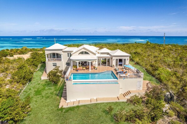 A large, modern house with a swimming pool, surrounded by lush greenery and overlooking a clear blue ocean.