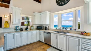 A kitchen with white cabinets, a granite countertop, a dishwasher, a microwave, and a window with a view of the ocean.