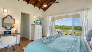 A bedroom with a bed, a desk with a laptop, a dresser, and a view of the landscape through large windows. - Villa Okeanos Anguilla (Shoal Bay Village)