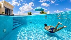 A swimming pool with clear blue water, a person swimming, and a building with a blue umbrella in the background.