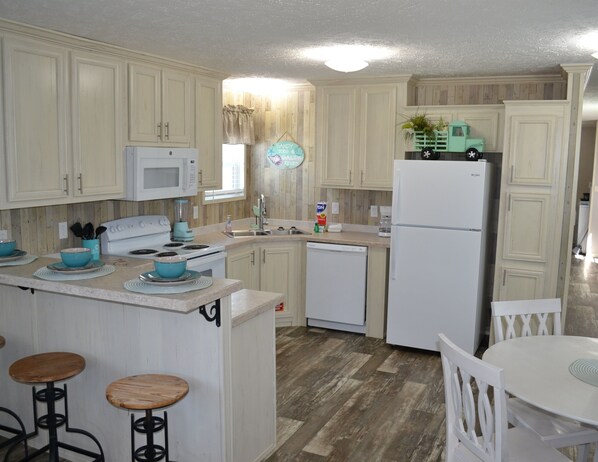 A kitchen with white cabinets, a microwave, a stove, a sink, and a refrigerator. There are stools and a table with chairs. - Sum Beaches Paradise Cottage 2 (Navarre)