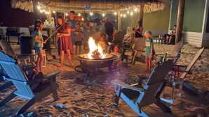 A beachside lounge with lounge chairs, a fire pit, and a thatched-roof structure.