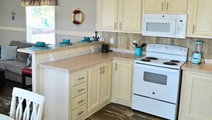 A compact kitchen with a microwave, stove, and a blender on the countertop. There is a small island with a sink and a few bowls on it. The kitchen has light-colored cabinets and a wooden floor.