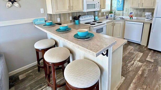 A kitchen with wooden cabinets, a central island with stools, a microwave, stove, and a dishwasher. - Sum Beaches Paradise Cottage 6 (Navarre)
