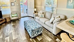 Un salon chaleureux avec un plancher en bois, un canapé beige, un pouf à motifs et un ventilateur de plafond.