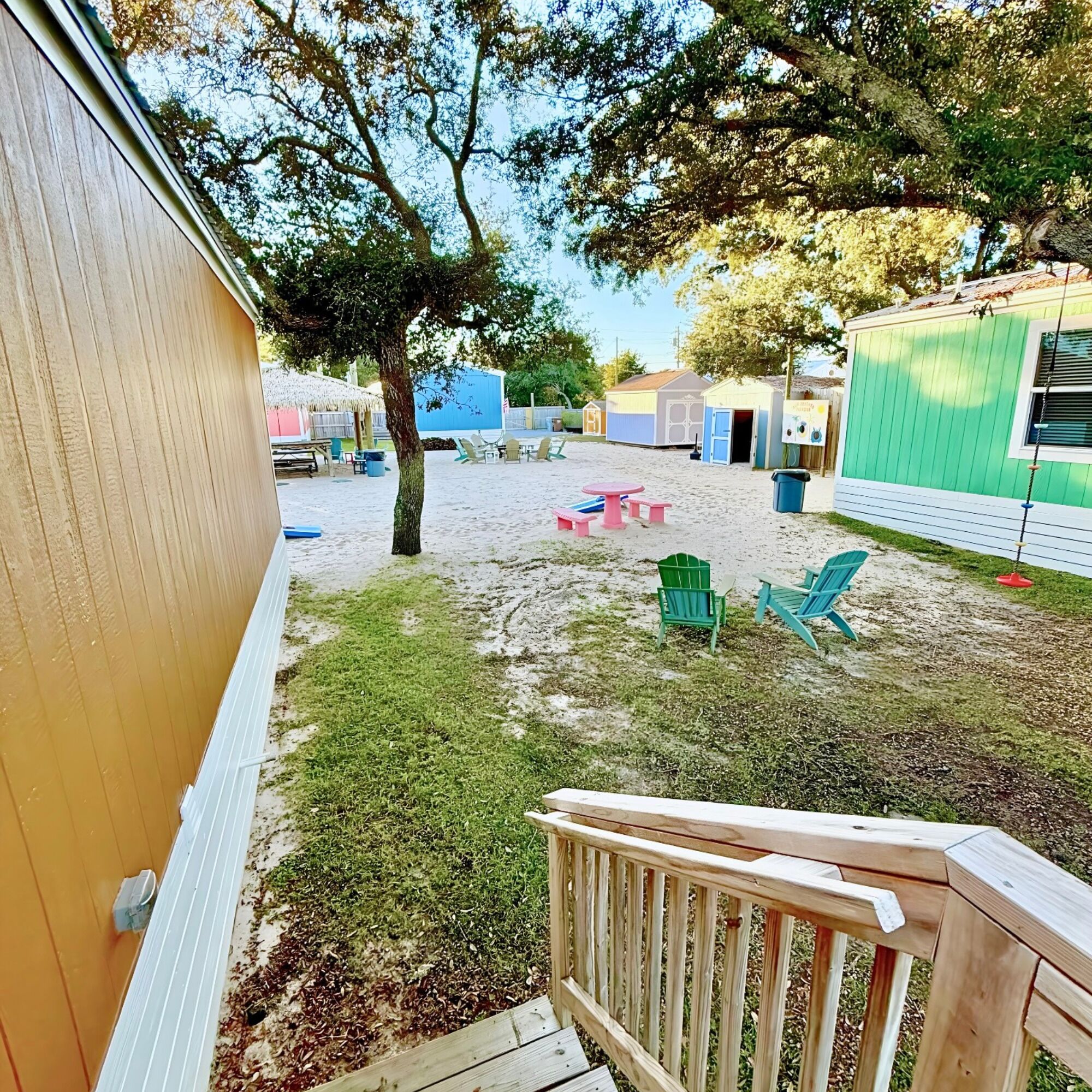 Un jardin avec une terrasse en bois, une table de pique-nique et des chaises vertes. On aperçoit de petites constructions et un arbre au fond.