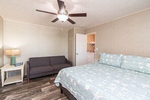 A bedroom with a bed, a gray sofa, a nightstand with a lamp, and a ceiling fan.