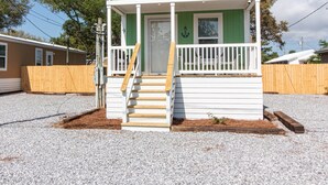 A small, single-story building with a green exterior and white trim, featuring a front porch with a wooden railing and a set of stairs leading to the door. - Sum Beaches Paradise Cottage 9 (Navarre)
