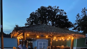 A beachside hut with a thatched roof, surrounded by beach chairs and a dining table. - Sum Beaches Paradise Cottage 9 (Navarre)