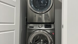Une machine à laver et un sèche-linge superposés dans une buanderie, avec un plancher en bois et des murs blancs.