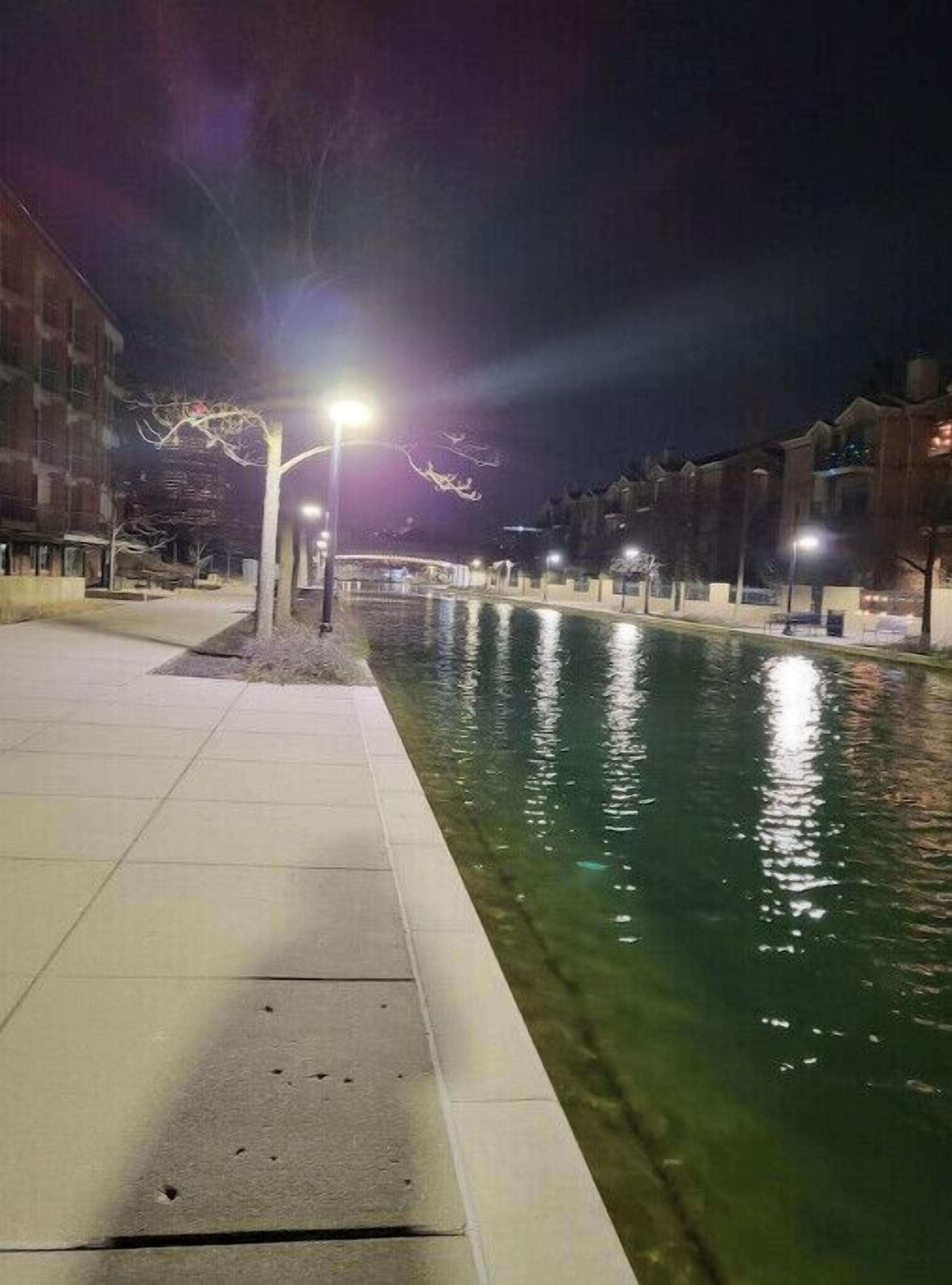 Ein Nachtbild eines Kanals mit Bürgersteig auf einer Seite und Gebäuden auf der anderen. Straßenlaternen säumen den Kanal, und das Wasser spiegelt die Lichter.