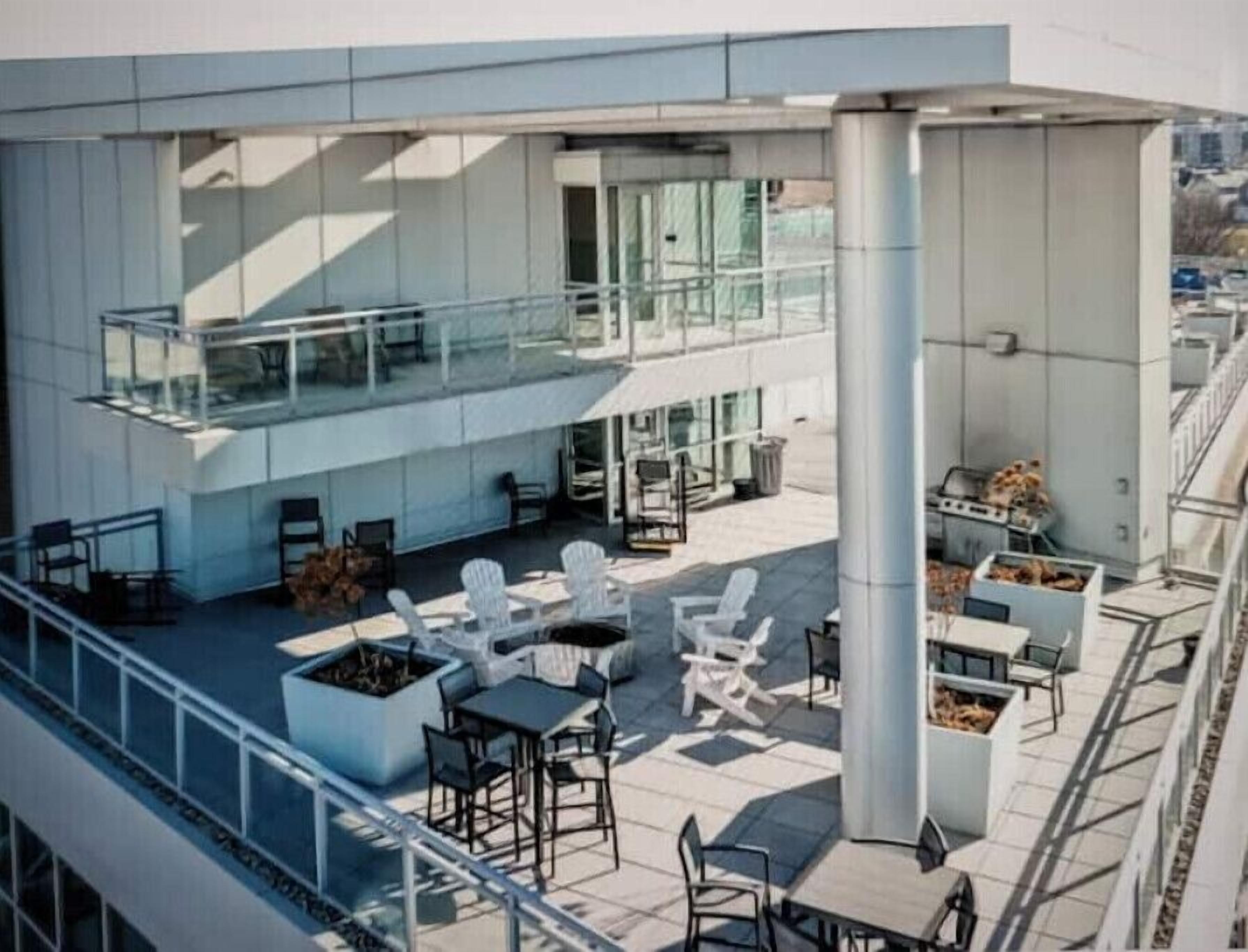 Un espace extérieur moderne, aménagé avec des tables et des chaises, doté d’une balustrade en verre et offrant une vue sur le paysage urbain.
