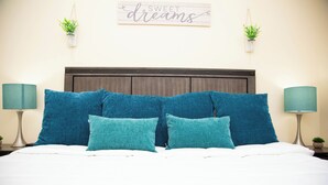 Un lit bien fait, agrémenté d’oreillers bleus et turquoise, d’une tête de lit en bois foncé et d’une inscription sur le mur qui dit : « Faites de beaux rêves ».