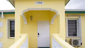 Un bâtiment jaune avec une porte blanche, le numéro « 115A » au-dessus de la porte, et un escalier avec des rampes blanches menant à l’entrée.