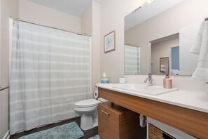A bathroom with a shower curtain, toilet, sink, mirror, and a towel hanging on the wall. - Sweet Tart Spot - Cozy TC Condo - Tart Trail - Balcony - Pets Welcome (Traverse City)