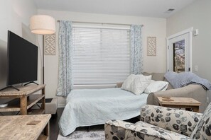 Un dormitorio con cama, televisión, sofá, una mesita y una ventana con persianas.
