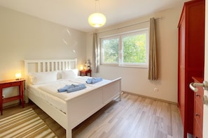 A bedroom with a bed, a nightstand, a dresser, a window with curtains, and a wooden floor. - Relaxed vacation in the Strandstraße: Your dream home by the sea! - (Loddin)