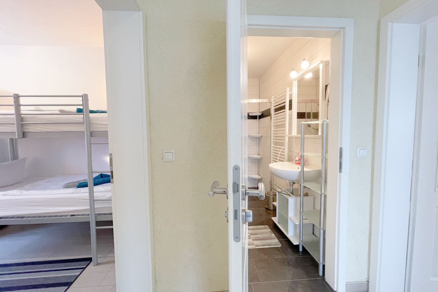 A room with bunk beds, a bathroom with a sink and mirror, and a door leading to another room.